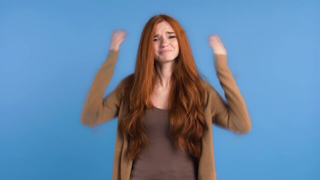Redheaded woman in casual outfit. She raised her hands up like asking why, looking very upset and annoyed while posing on blue background. Close up