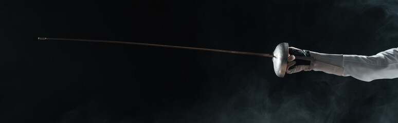 Panoramic orientation of fencer in glove holding rapier on black background with smoke