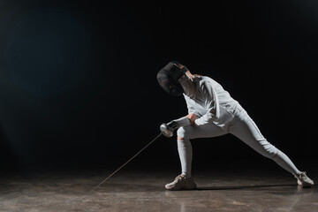 Fototapeta na wymiar Fencer in fencing mask and suit holding rapier and doing lunge on black background
