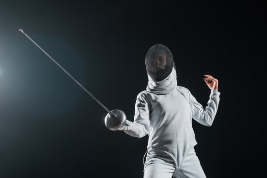 Low angle view of fencer exercising with rapier on black background with lighting