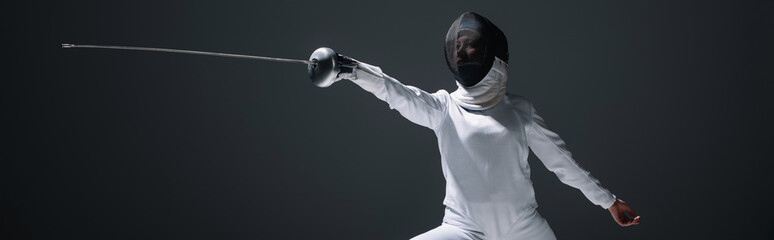 Panoramic shot of fencer in fencing suit and mask exercising isolated on black