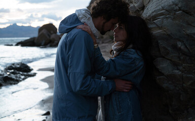 Two lovers tenderly look into each other's eyes nestled against the rock on the beach during the sunset.