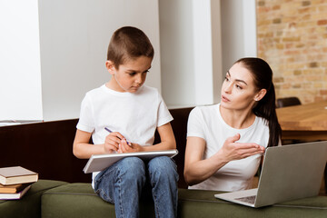 Fototapeta premium Mother point with hand at laptop to son holding color pencil near books on couch