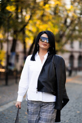 Fototapeta premium Autumn fashion outdoor. The girl in fashionable stylish white shirt , pants, jacket and sunglasses, autumnal lifestyle on the background of blurry yellow-green trees in the park. Vertical