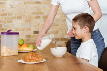 Mother pouring milk in bowl with cereals near smiling son at kitchen table