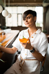 Beautiful woman with short brunette hair in white clothes. Fashion street photography. Fashion model posing with aperol cocktail on wood background.