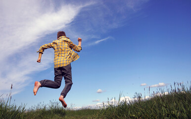 A teenager in a yellow shirt in a cage rejoices at summer and holidays and jumps into a field on green grass barefoot. Blue sky, copy space   