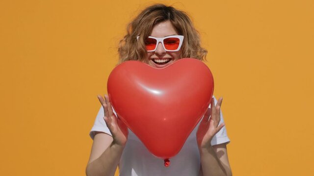 Portrait of happy woman with red heart balloon holding in her hands moving from side to side in front of her lowers down slow motion smiling at camera on yellow background in summer. Emotion. Flirt