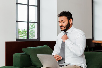 african american freelancer in suit touching tie near laptop