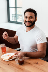 Fototapeta na wymiar happy african american man holding spoon near jar with sweet jam and toast bread on plate