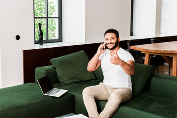 bearded african american man talking on smartphone and showing thumb up near laptop with blank screen