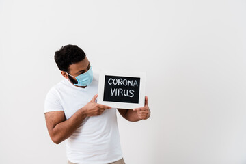 african american man in blue medical mask looking at chalkboard with coronavirus lettering near white wall