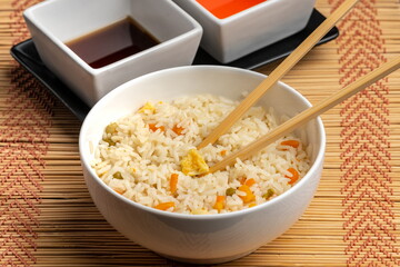 Chinese food bowl of rice with chopsticks and soy sauce and sweet and sour sauce on bamboo trivet and black background
