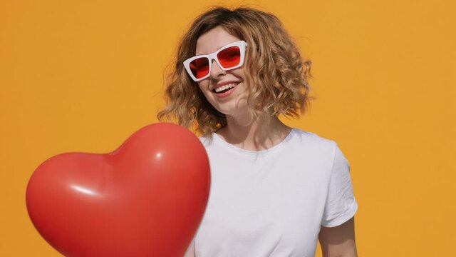 Portrait of happy woman smiling red heart balloon holds in her hands moving from side to side in front of herself at camera on yellow background in summer. Emotions. Positive. Flirt. Valentine. Love