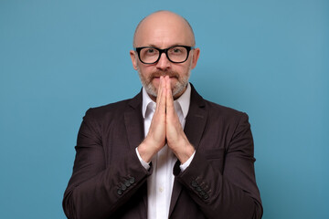 Miserable mature man in suit and glasses with begging expression keeps hands in praying gesture....
