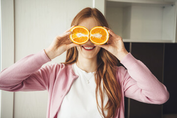 Red haired caucasian woman smiling at camera while cover her eyes with oranges in the kitchen