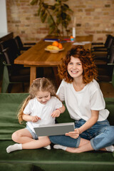 Fototapeta premium cheerful redhead mother holding laptop near cute daughter in living room