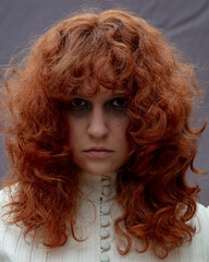 Fototapeta premium A portrait of a young woman with long curly, red hair, wearing a vintage dress and looking sad and serious.