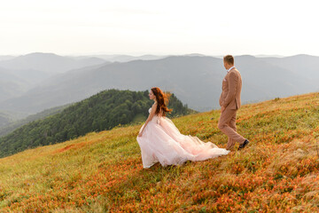 The bride and groom go next to each other. Sunset. Wedding photo on a background of autumn mountains. A strong wind inflates hair and dress.