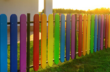 Wooden fence for kid-friendly white house consisting of strips of red, orange, yellow, green, blue and violet panels. Rural view at white house with sunshine light. Amazing weekend staycations at home