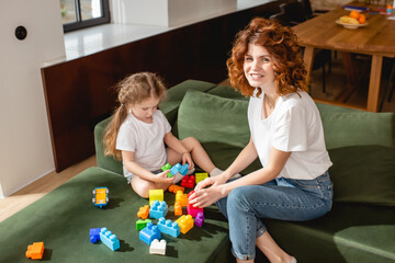 Obraz premium curly mother smiling near daughter playing building blocks in living room