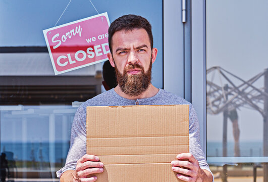 Unemployed Guy With Empty Signboad Near Closed Door Office Or Cafe. Unemployment During Coronavirus Covid-19 Crisis, Concept Of Job Loss