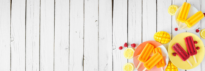 Selection of colorful fruit popsicles. Top down view table scene. Corner border on a white wood banner background with copy space.
