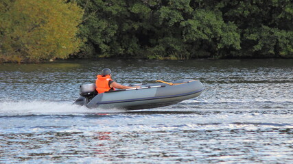 Grey Inflatable outboard motor boat with racer in brighy orange lifejacket and helmet fast glide floats on water Race at river on shore trees background at Sunny summer day, outdoor sport competition