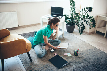 Caucasian woman with eyeglasses studying on the floor from book and listening to music through earphones