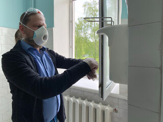 A man in a respirator and medical gloves treats his hands with an antiseptic. A man clicks on a dispenser for an antiseptic in a hospital after receiving a doctor.