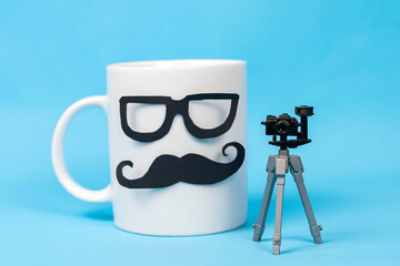 White mug with glasses and mustache and a little photography camera with tripod on blue background