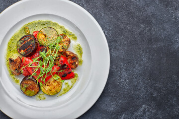 Grilled baked vegetables, eggplant, zucchini, bell pepper, champignon mushrooms, on a white plate against a dark background, with microgreen