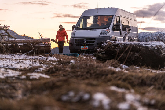 Man by a camper van carrying skis in a rocky wintry landscape.