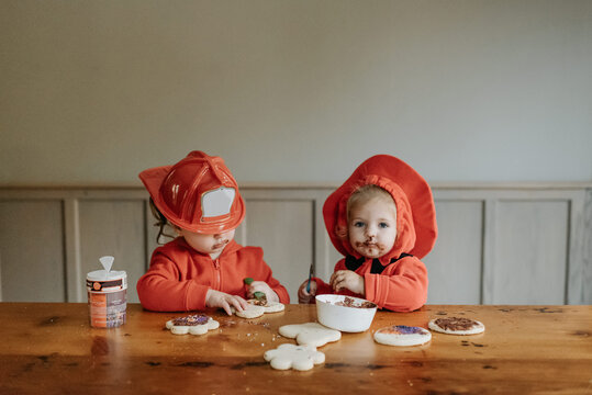 Two children dressed as firefighters at a table decorating  cookies with chocolates and sprinkles.