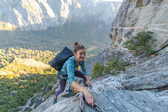 A woman climbing up a mountain with a valley full of pine trees below.