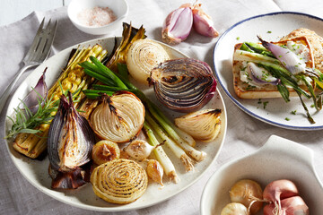 Plates and dishes with a variety of roasted onions, a selection of onions on toast and uncooked shallots in a bowl.