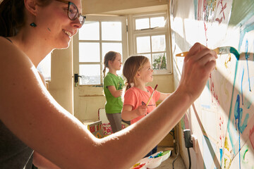 Smiling woman and two girls indoors during Corona virus crisis, painting on a wall.