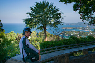 The young man sits has a rest on the observation, survey deck. A tourist in a helmet and with a backpack poses on vacation. The Andamand Sea of Phuket Island, Thailand.