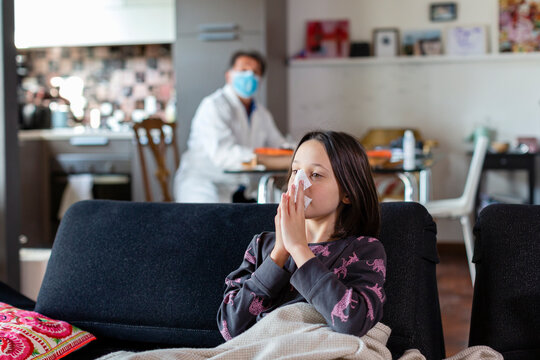 Girl sitting on a sofa blowing her nose, and a doctor seated in a face mask and white coat at a distance.