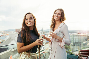 Two women dressed to go out for the evening standing on a balcony overlooking a city holding glasses of champagne.