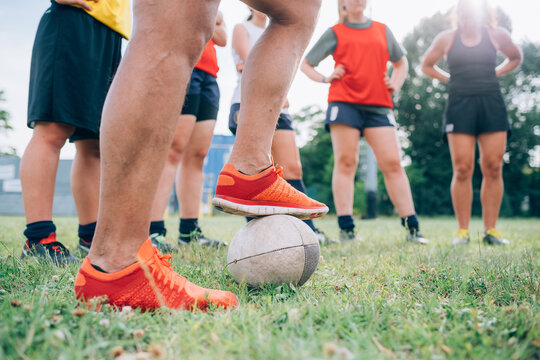 The legs and feet of a group of women standing on a training pitch one with a foot on a rugby ball.
