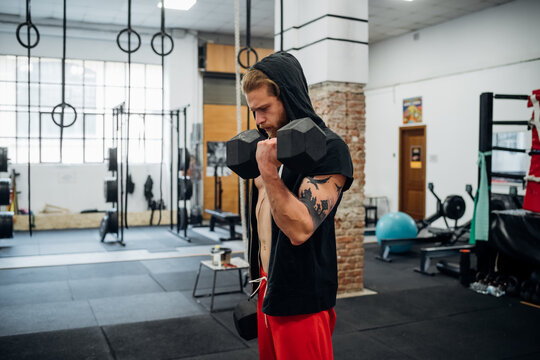 A man standing in a gym lifting a large dumbell with his left arm.