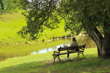 A woman sits on a wooden bench near the water. A lonely woman is in a public park.