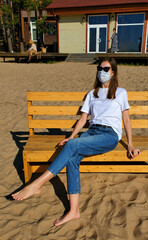 A 30-year-old girl of European appearance in a protective medical mask, sunglasses, a white T-shirt and jeans sits on a wooden bench on a sandy beach. Quarantine rest, isolation, lonely masked girl 