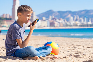Teenager boy using a smart phone voice recognition function online, using cell phone to send a voice message. Kid sitting on Benidorm city beach