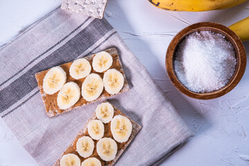 Healthy snack with Swedish rye crispbread, peanut butter and banana's slices on a cloth and a wooden bowl with grated coconut. Top view. Flat lay.