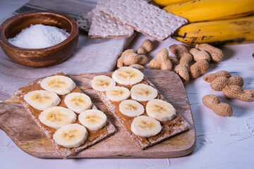 Crispbread with peanut butter and banana on a wooden table and other ingredients in background. Healthy snack. Close up view.