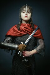 Gorgeous young woman in knight armour and steel chainmaille holding sword on dark background. Strong and sensual woman with perfect skin and with natural makeup.
