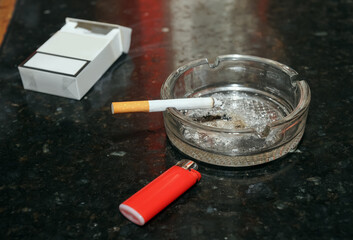 Cigarro encendido en un cenicero junto a un encendedor y un paquete de tabaco.