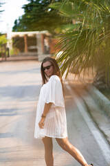 Young beautiful woman in white dress and sunglasses turns around on the road with palms. The concept of joy, ease and freedom during the vacation. The girl is enjoying the rest. Fashion